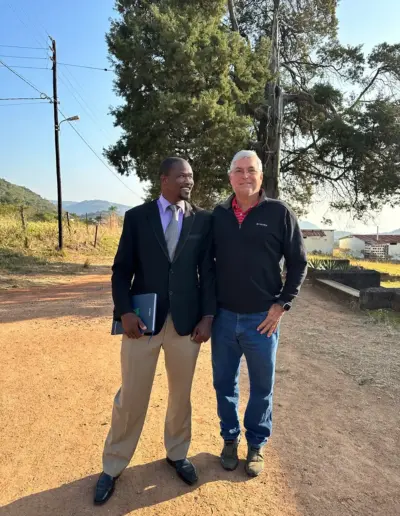 Two men standing on rural dirt road with trees and hills in background.