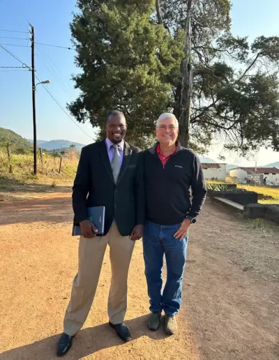 Two men walking together on a rural dirt road with trees and fields in background.
